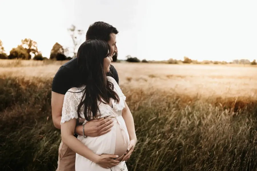 Paar im hohen Gras, die Hände um den Babybauch gelegt, die Sonne im Rücken