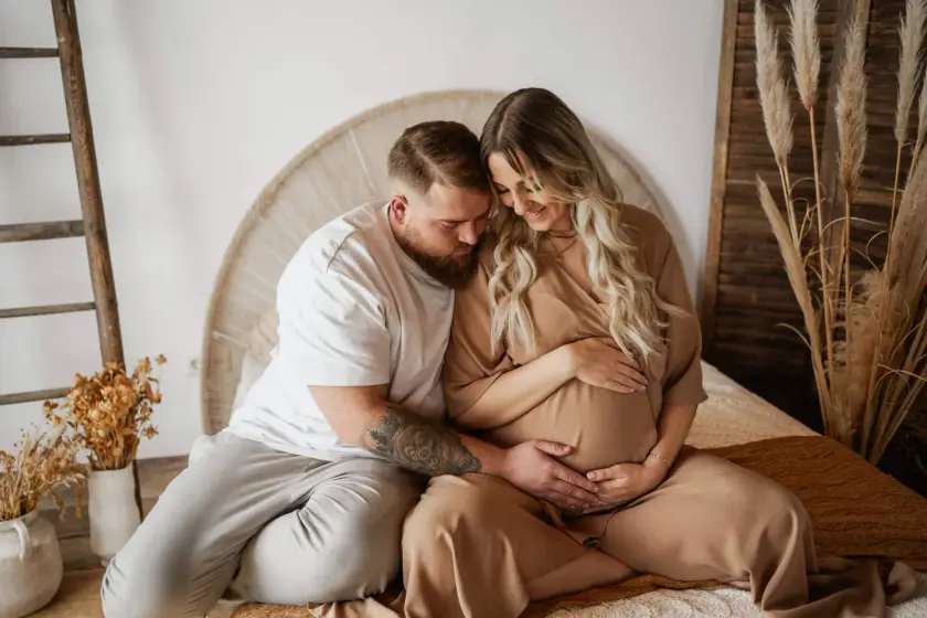 Werdende Eltern sitzen im hellen Studio auf dem Boden, sie lehnt sich an ihn, beide halten den Babybauch