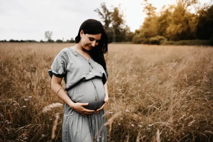 Schwangere Frau im grauen Kleid steht allein im hohen, goldenen Gras und blickt lächelnd auf ihren Bauch
