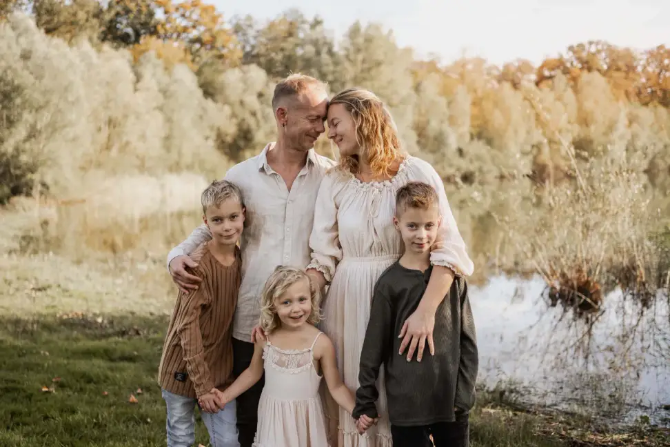 Familie mit drei Kindern steht eng zusammen am Seeufer, die Eltern schauen sich lachend an