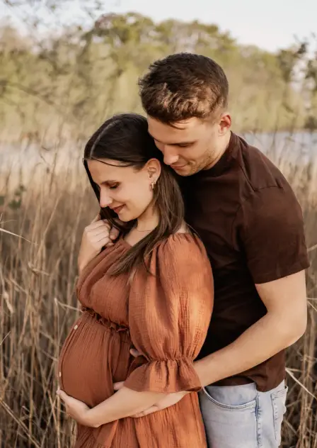 Werdende Eltern im Schilf, sie im rostroten Kleid, er legt von hinten die Arme um sie und ihren Bauch