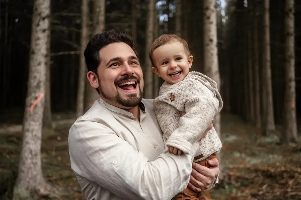 Ein Vater lacht mit weit offenem Mund, sein Baby auf dem Arm lacht mit, im Hintergrund ein Tannenwald