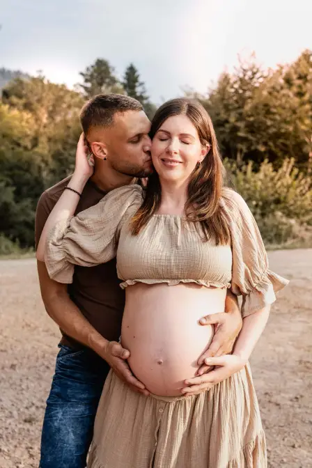Er küsst ihre Wange und hält ihren nackten Babybauch, sie schließt lächelnd die Augen, im Hintergrund Bäume