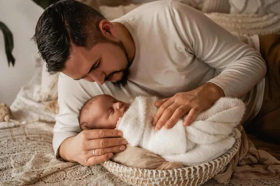 Ein Vater beugt sich über sein Neugeborenes im Körbchen und legt behutsam die Hände um es