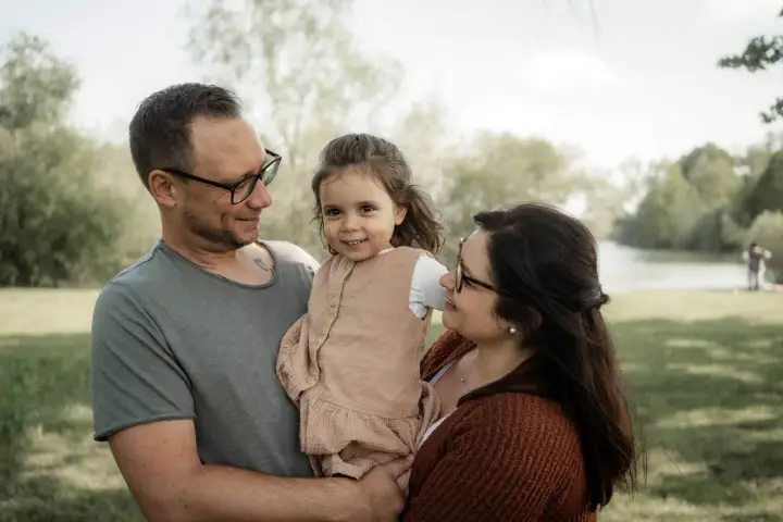 Eltern halten ihre kleine Tochter zwischen sich auf dem Arm, alle drei lachen, im Hintergrund ein See