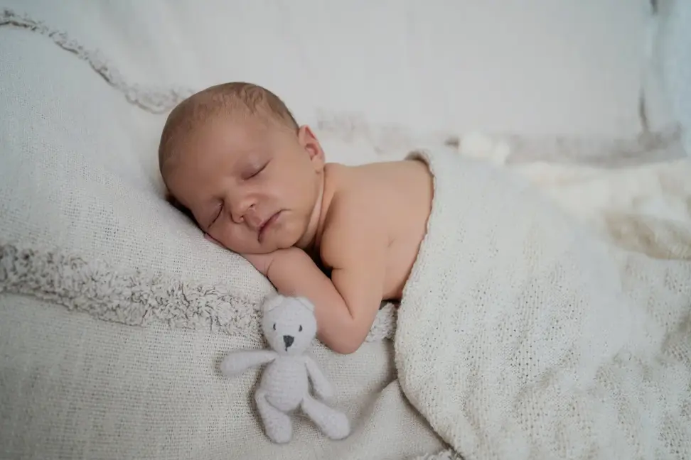 Neugeborenes schläft auf einer weißen Decke, neben ihm ein kleiner Stoffteddy