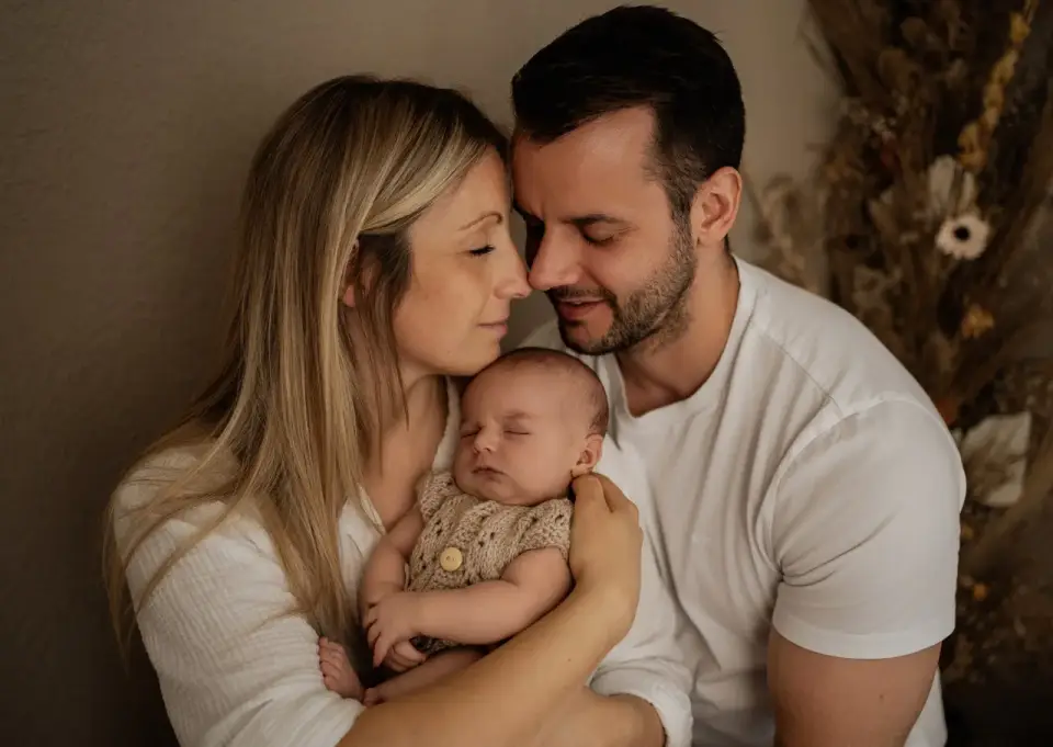 Neugeborenenshooting im Studio: Mutter und Vater lehnen Stirn an Stirn und halten ihr schlafendes Baby im Strickbody zwischen sich
