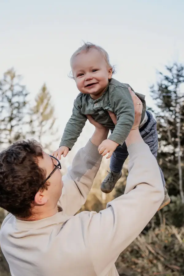 Ein Vater hebt sein lachendes Baby hoch in den Himmel, dahinter Bäume
