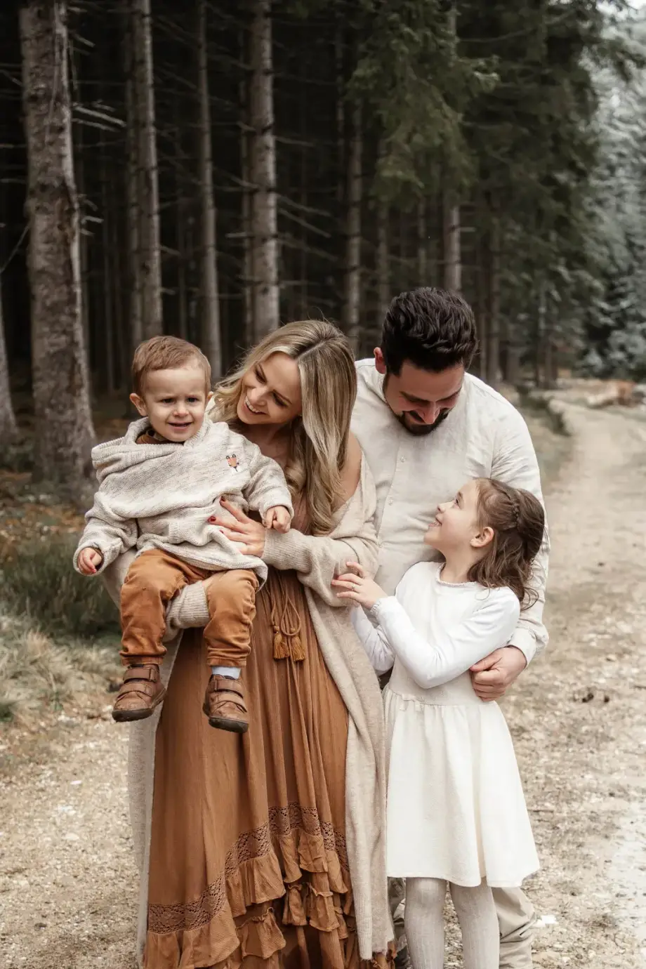Familie auf einem Waldweg zwischen hohen Tannen, die Mama trägt das Kleinkind, der Papa beugt sich lachend zur Tochter