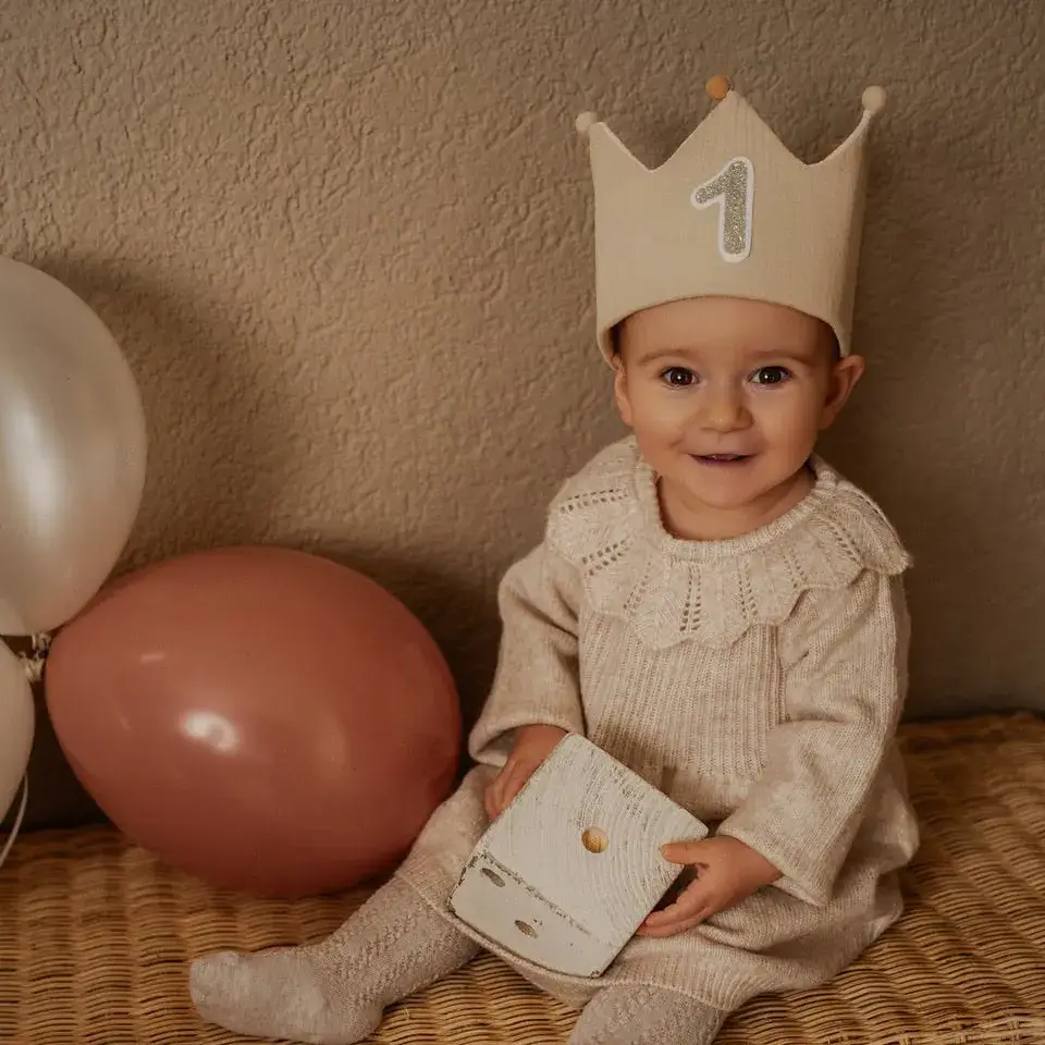 Ein einjähriges Kind mit Stoffkrone und der Zahl 1 sitzt lächelnd vor Luftballons, ein kleines Buch auf dem Schoß