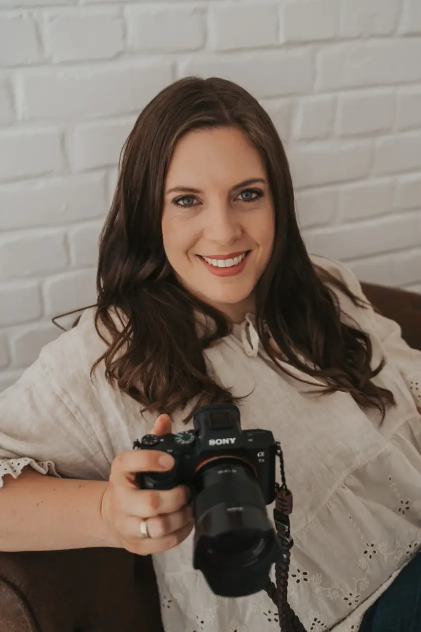 Marion Leistler, lächelnd, in einer hellen Leinenbluse, ihre Kamera in der Hand, vor einer weißen Backsteinwand