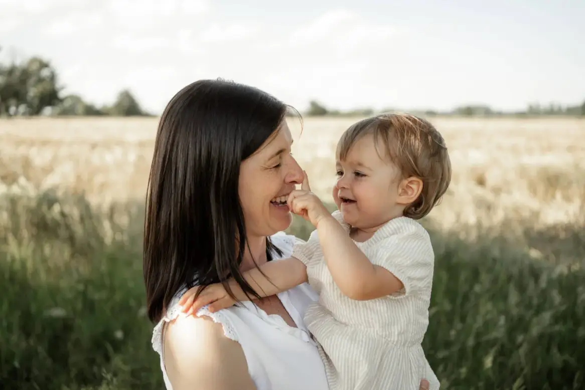 Eine Mutter hält ihre kleine Tochter auf der Wiese, beide lachen, das Mädchen greift nach ihrer Nase