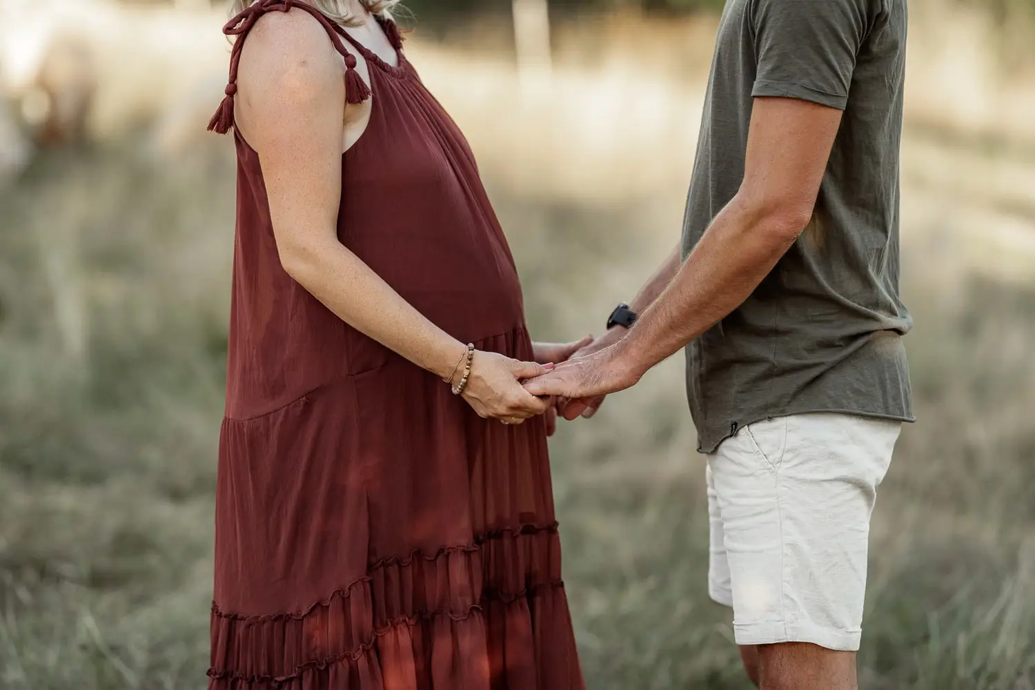 Nahaufnahme: Er hält die Hände seiner schwangeren Frau im weinroten Kleid, auf Höhe ihres Babybauchs, im Hintergrund eine Sommerwiese