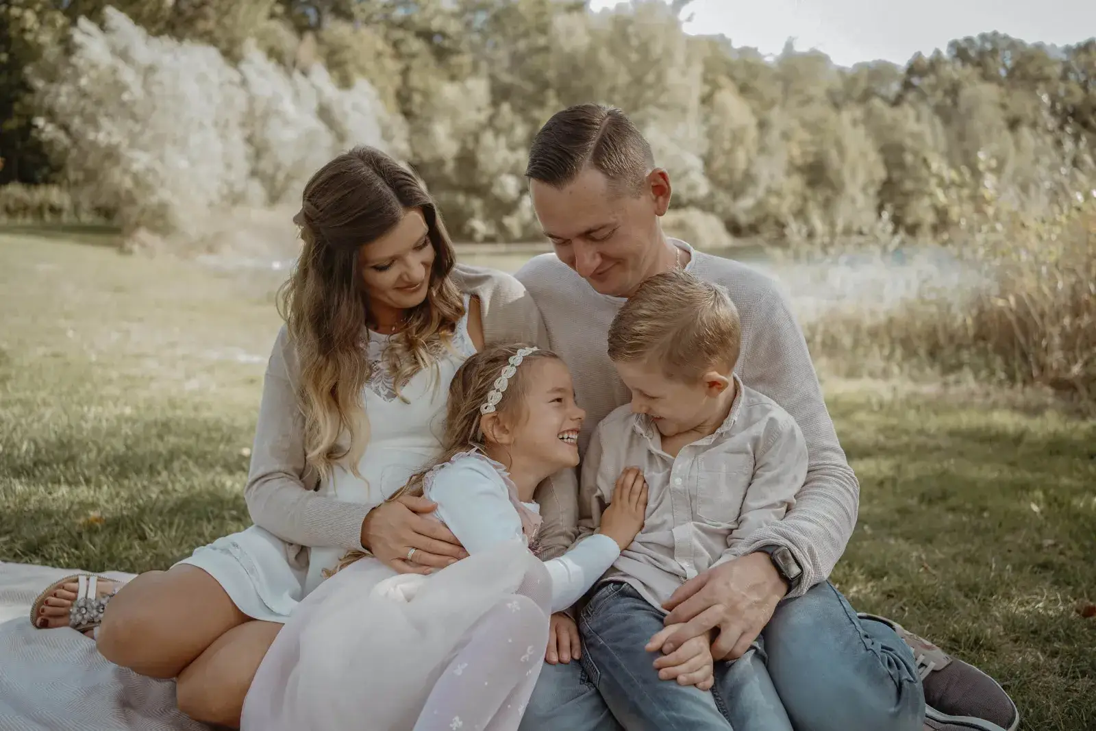 Familie sitzt eng beieinander auf einer Sommerwiese, die Mama hält die Tochter, der Papa umarmt den Sohn, im Hintergrund ein See