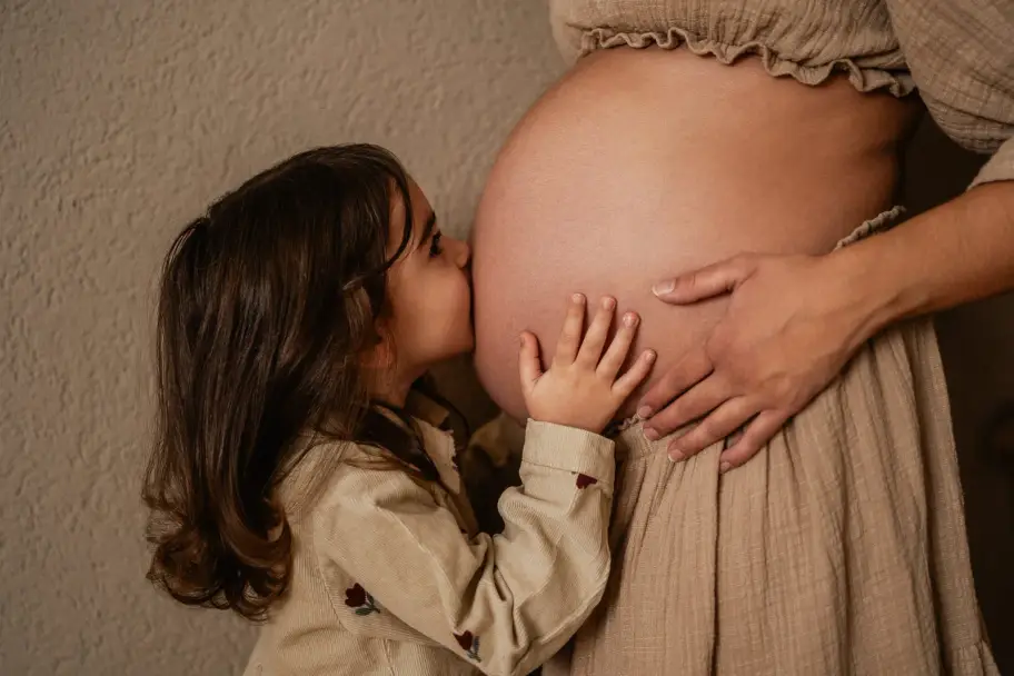Ein kleines Mädchen küsst den nackten Babybauch ihrer Mama, die ihre Hände um den Bauch legt