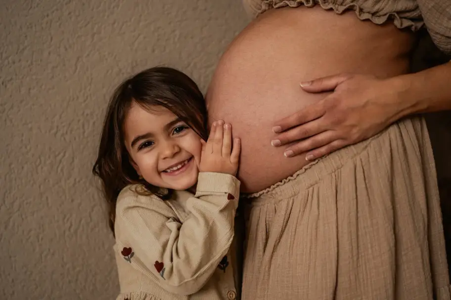 Das Mädchen legt lachend ihre Wange an den Babybauch und lauscht, die Hände der Mama liegen darüber