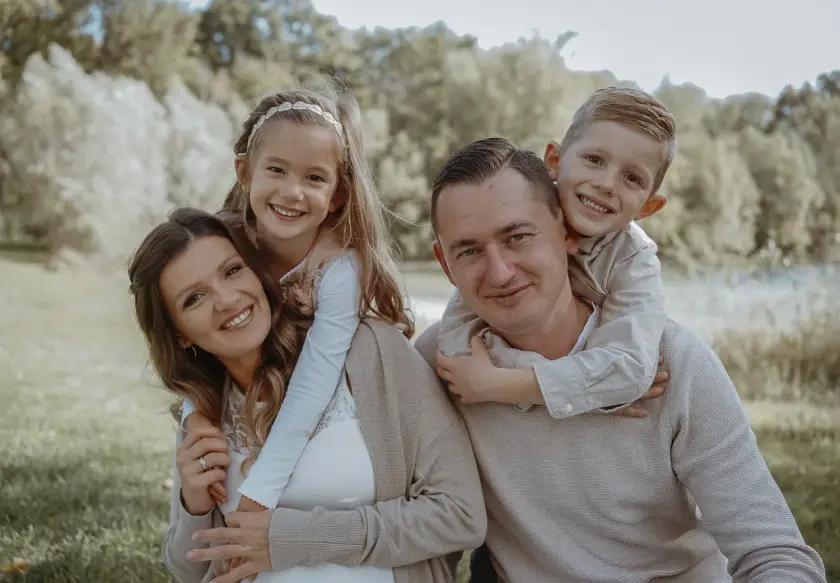 Familie mit zwei Kindern auf einer Wiese, die Tochter umarmt die Mama von hinten, der Sohn klettert dem Papa auf den Rücken, alle lachen