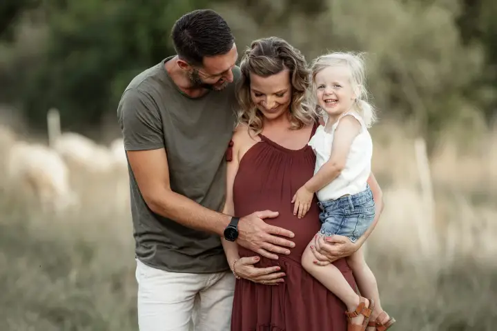 Werdende Eltern mit ihrer kleinen Tochter auf dem Arm, sie im weinroten Kleid, er legt die Hand auf ihren Bauch