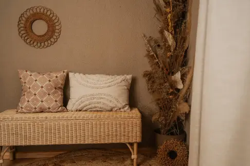 Rattanbank mit Kissen vor einer warm getönten Wand, daneben ein Gesteck aus Trockenblumen und ein runder Spiegel