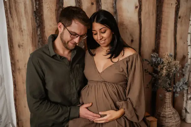 Werdende Eltern vor einer Holzwand, sie im braunen Kleid, er legt die Hände auf ihren Bauch, beide schauen hinunter