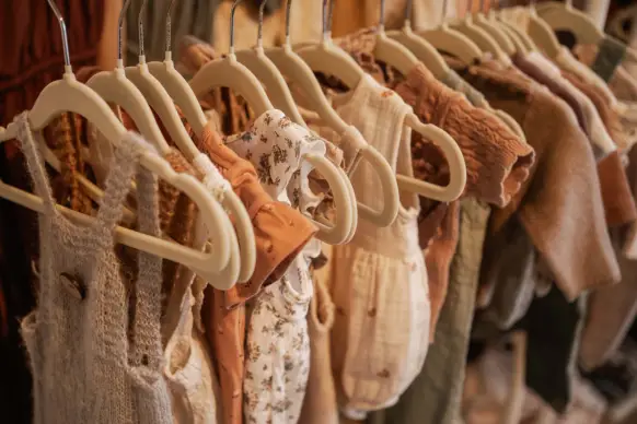 Eine Kleiderstange voller kleiner Babyoutfits in Erdtönen, Strick und Leinen auf hellen Holzbügeln