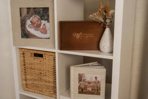 Weißes Regal mit gerahmtem Babyfoto, einer Holzbox mit Logo, einem Korb und einem Fotoalbum