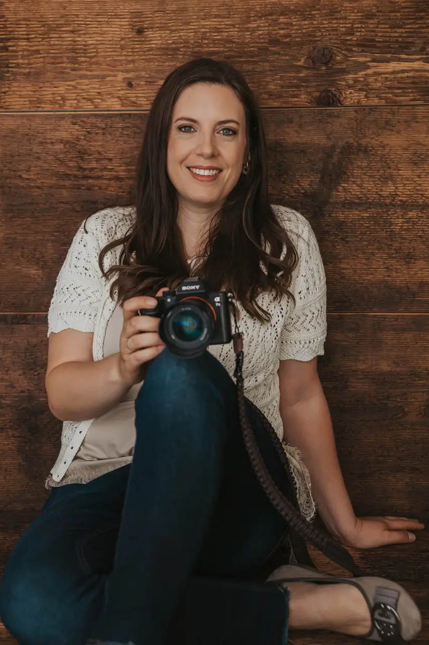 Marion Leistler sitzt lächelnd vor einer dunklen Holzwand, im hellen Strickcardigan und Jeans, die Kamera auf dem Knie