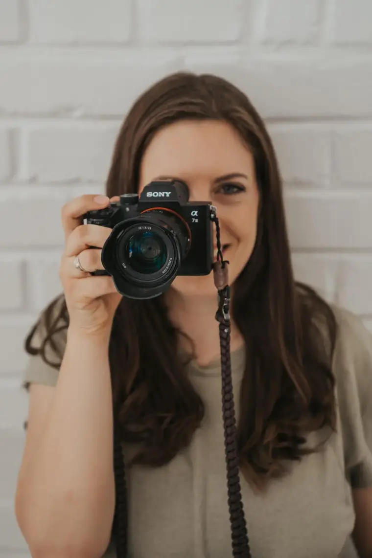 Marion Leistler hält ihre Kamera vors Gesicht und fotografiert direkt in Richtung Betrachter, dahinter eine weiße Backsteinwand