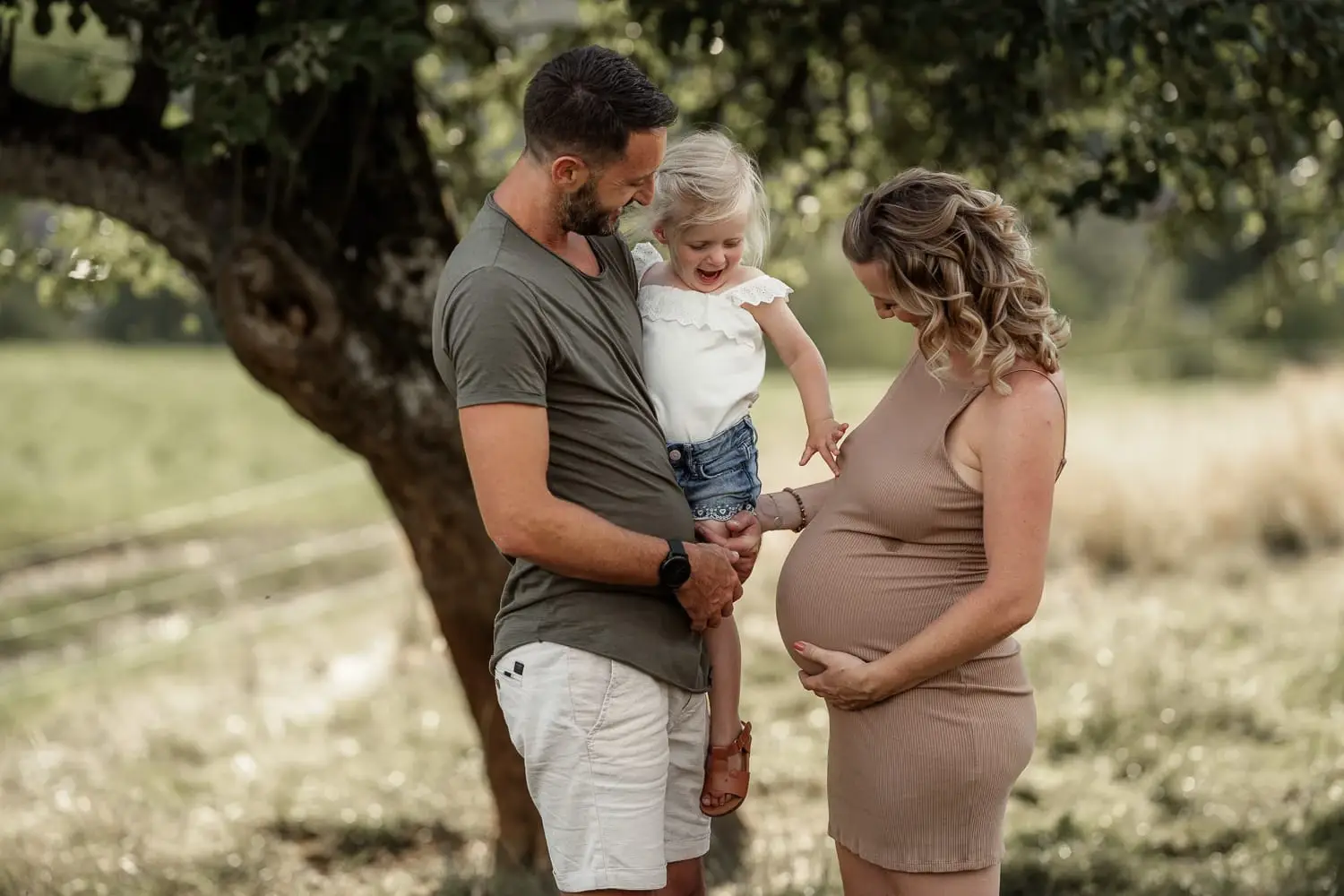 Eine schwangere Mama steht unter einem Baum, der Papa hält die kleine Tochter auf dem Arm und legt die Hand auf den Babybauch