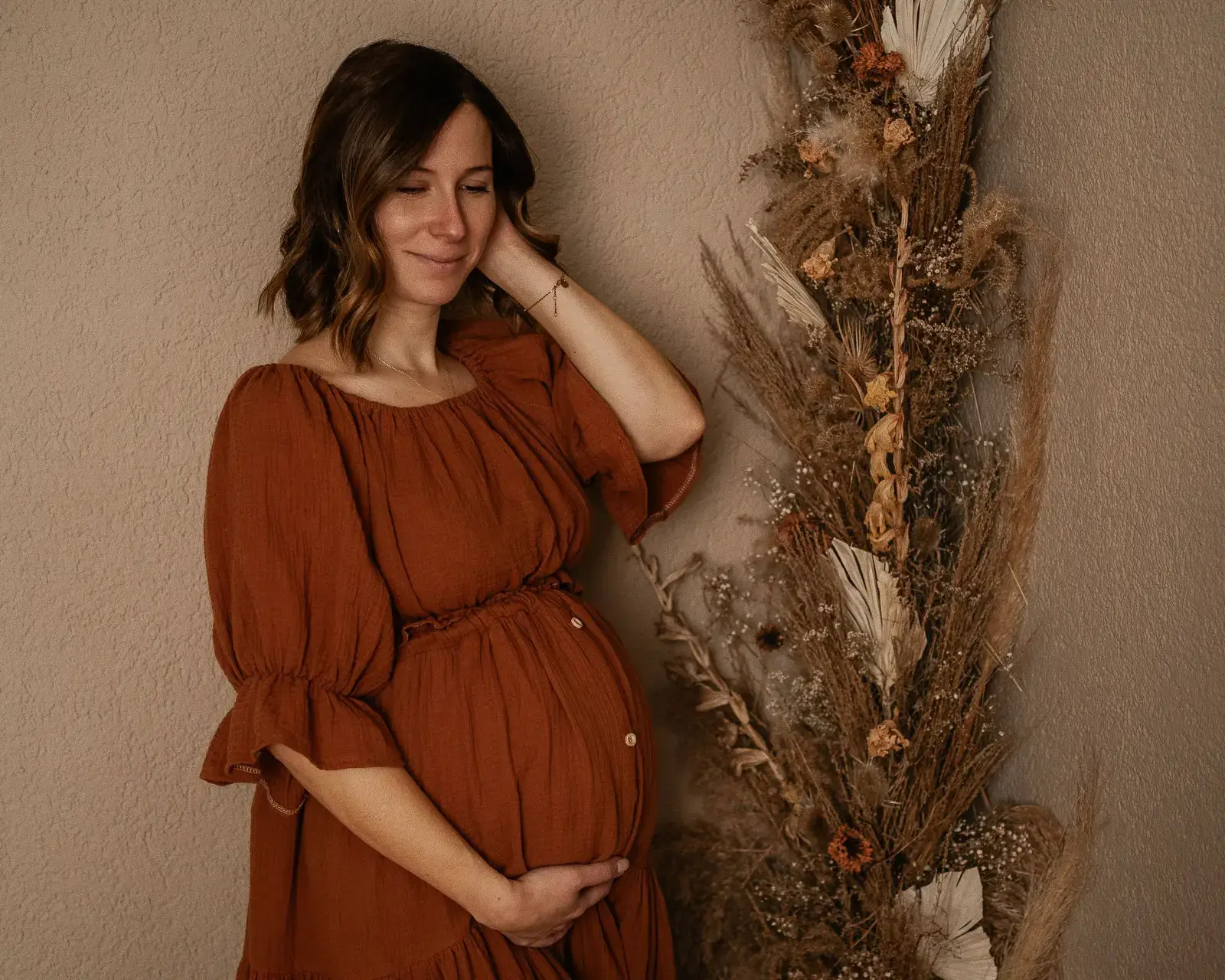 Eine schwangere Frau im rostroten Kleid steht lächelnd im Studio, eine Hand am Bauch, daneben Trockenblumen